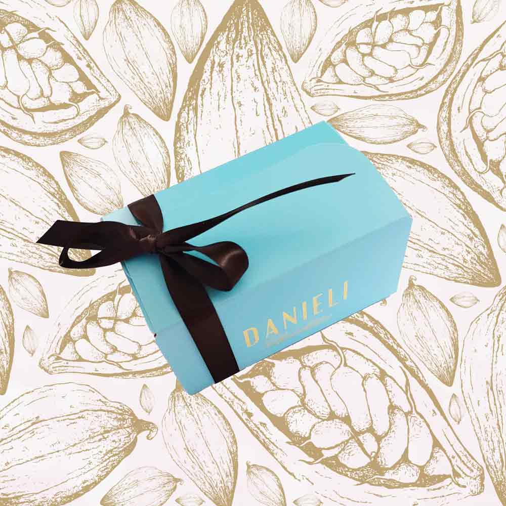 Blue gift box with a black ribbon on a background with cocoa bean illustrations, featuring the brand 'DANIELI'.